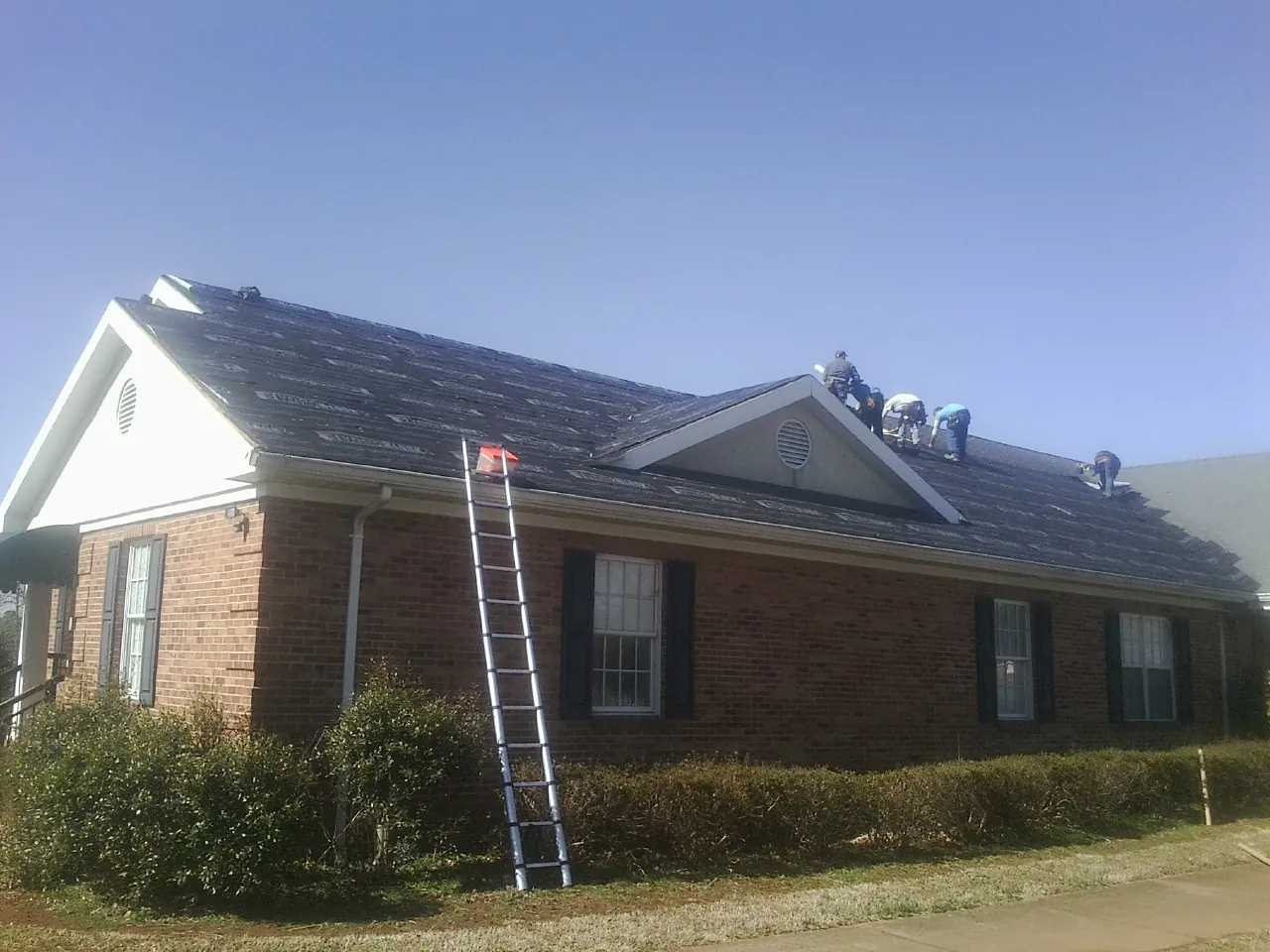 Crew tearing shingles off a brick ranch roof with a ladder set — Metal Roof Repair work by our crew in Rochert